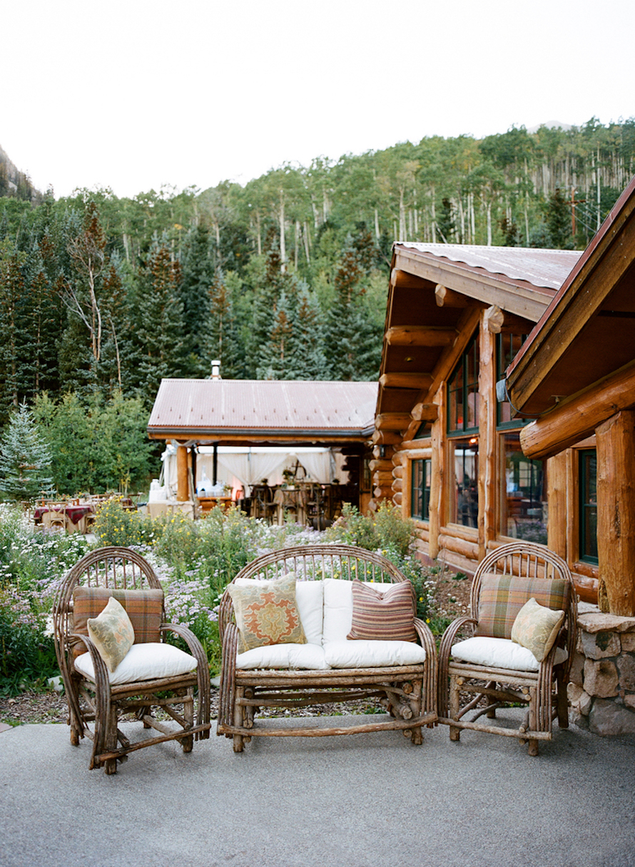 Summer evening on the Pine Creek Cookhouse patio &mdash; willow lounge chairs, wildflowers, and the log cabin beyond.